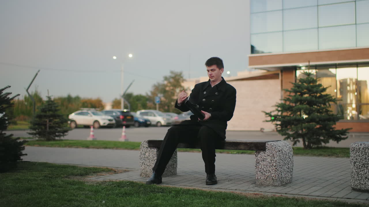 검은색 슈트를 입은 사업가가 벤치에 다가와, 현대적인 건물과 배경에 주차된 자동차와 함께 야외 설정에서 브리프케이스를 열고, 전문, 도시, 사무실 직원이 휴식을 취합니다.