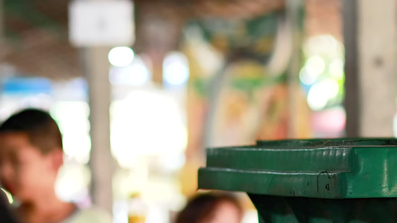 A person places a yellow box into a green waste bin at a market setting.