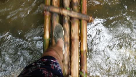 a pov shot of a young man walking over a bamboo bridge above a small stream wearing his sneakers and shorts