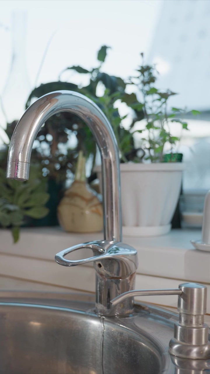 Close up of chrome kitchen faucet with water flow. Faucet is shiny and metallic with curved spout and lever handle. Scene is kitchen sink area with natural light