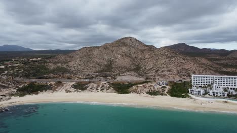 Toma-Aérea-Hacia-Atrás-Del-Hermoso-Paisaje-Costero-En-Los-Cabos-Con-Vista-A-La-Playa-De-Arena,-Una-Concurrida-Carretera-Costera-Y-El-Mar-Turquesa-Y-El-Renombrado-Hotel-Marquis-Durante-Unas-Vacaciones-En-México