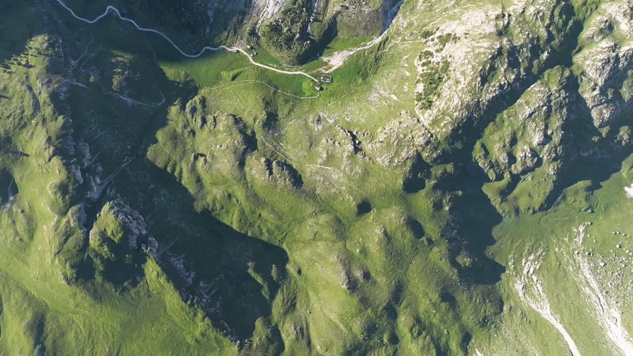 A bird's-eye view showcases the contrasting landscapes of the Dolomites: a meandering gravel street leading to a secluded mountain hut, juxtaposed against the raw, imposing mountain faces