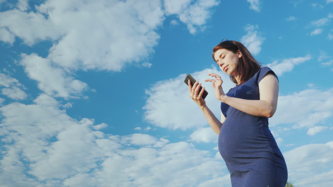 беременная женщина пользуется смартфоном на фоне голубого неба нижний угол обзора
