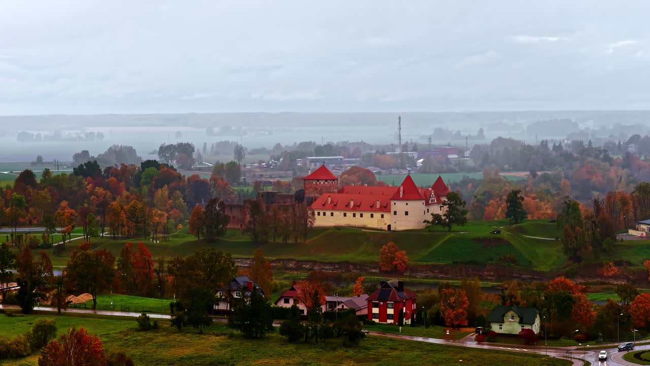 A misty overcast sky frames a red-roofed castle on a hill, surrounded by vibrant fall trees and a quiet village below