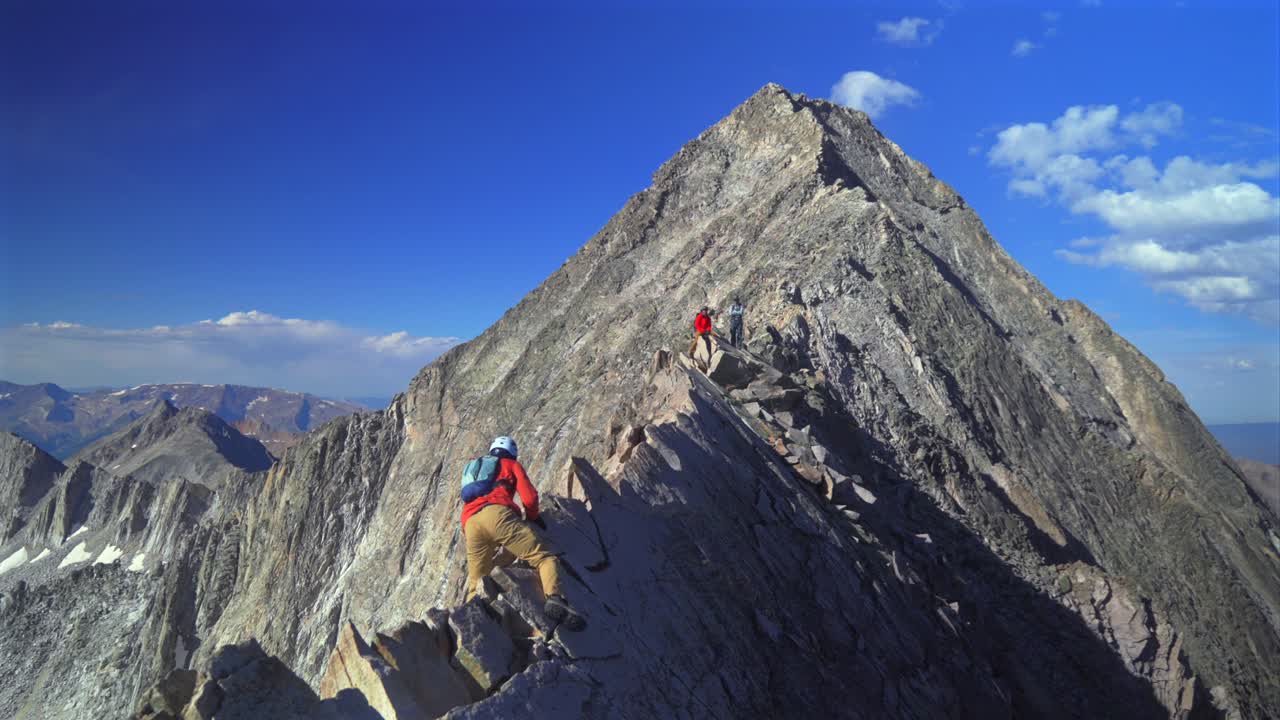 People hiking Knifes Edge North East Ridge trail Capitol Peak Wilderness Trail scrambling rock climbing Rocky Mountains Colorado 14er mountainside boulders summer blue sky morning sunny rugged terrain