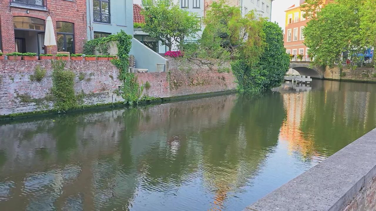 Naturskøn kanal omkranset af historisk murstensarkitektur, en buet stenbro, frodige grønne træer, efeu-beklædte facader, blomsterkasser og fredfyldte vandrefleksioner i en charmerende europæisk by.