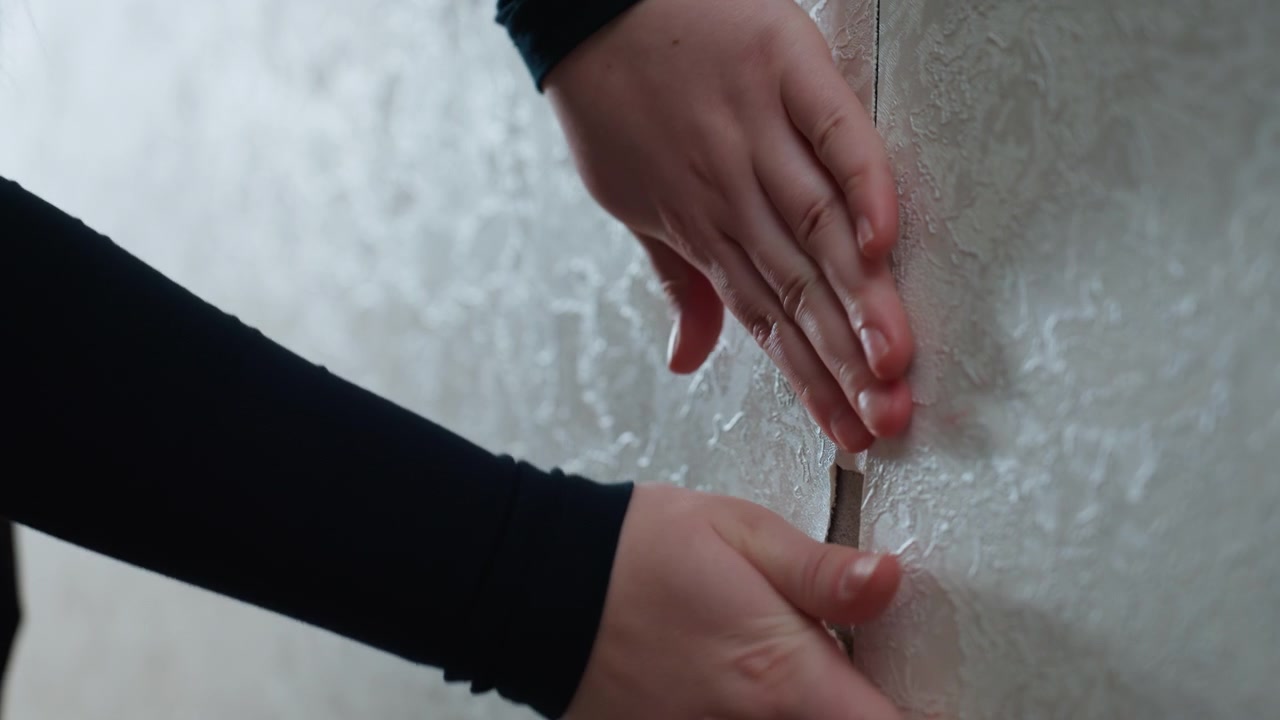Homeowner peels wallpaper from wall, careful hands expose seam and damaged plaster in bright new apartment during moving project tactile closeup, natural daylight, planning for repair and decorating