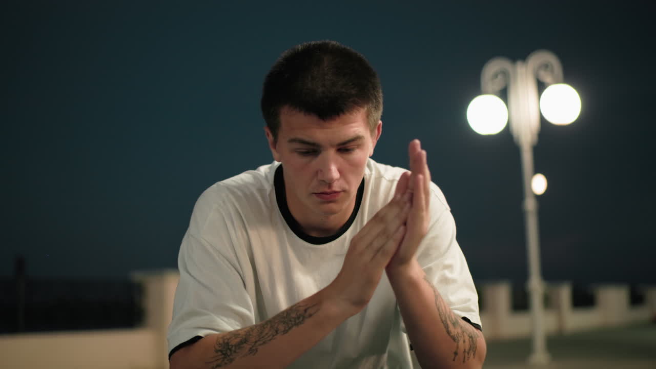 색 폴로 옷을 입은 젊은이가 손으로 얼굴을 고 손을 는 동안 손을 제거하고, 조용한 도시 환경에서 반사와 감정을 표현하는 배경에 streetlamps를 조명합니다.