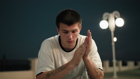 joven en polo blanco se cubre la cara con las manos y las quita mientras se frotan pensativamente, lámparas de la calle iluminadas en el fondo, expresando reflexión y emoción en un entorno urbano tranquilo