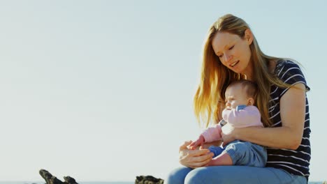 front view of caucasian baby sitting on mid-adult mothers lap at beach on a sunny day 4k