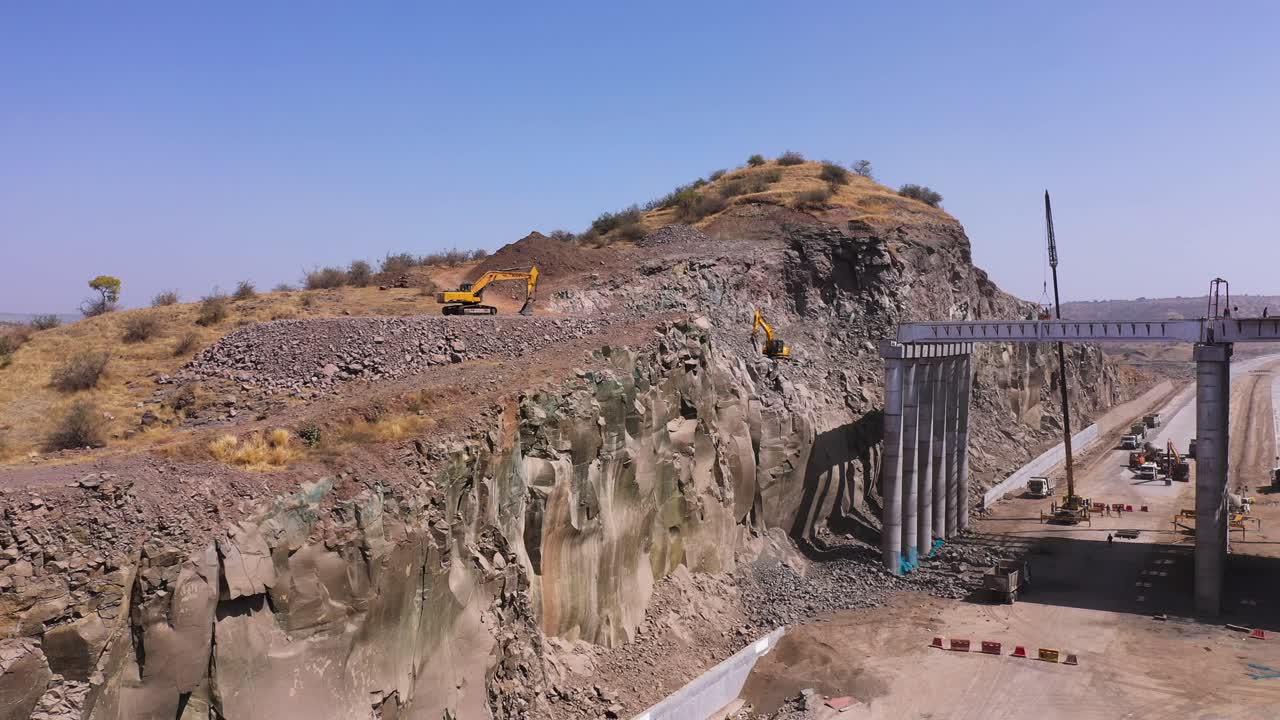 Aerial Pull Back Shot Of Two Excavator Trucks Beside A Mountain Cliff, Digging Through The Rocks In Relation To Pipeline Construction