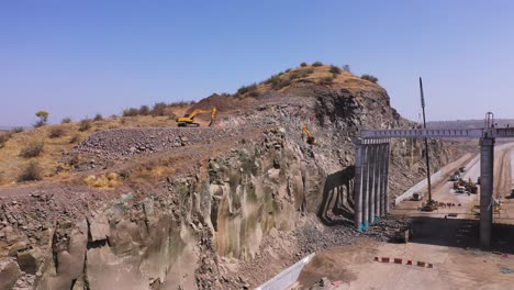 Aerial-Pull-Back-Shot-Of-Two-Excavator-Trucks-Beside-A-Mountain-Cliff,-Digging-Through-The-Rocks-In-Relation-To-Pipeline-Construction