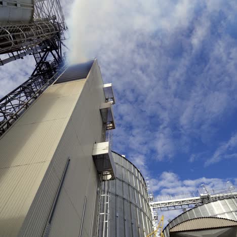 grain elevators against blue sky. thick white smoke releases from industrial building. modern complex for agribusiness. view from below.