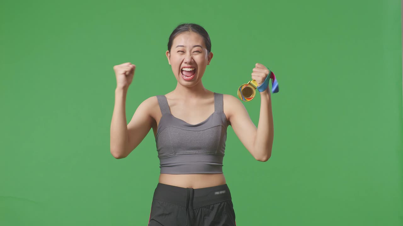 아시아 여성은 금메달을 바라보며 울부고 골을 치며 스튜디오의 녹색 스크린 배경에서 첫 번째 우승자로 우승을 축하합니다.