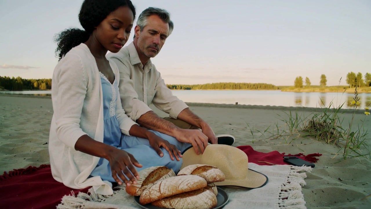 Lifting man placing hat on blanket at shore, woman in blue dress arranging bread for picnic