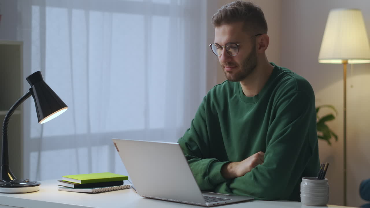 online communication by video chat young man dressed green sweatshirt and glasses is looking at display of notebook and nodding head portrait shot in living room
