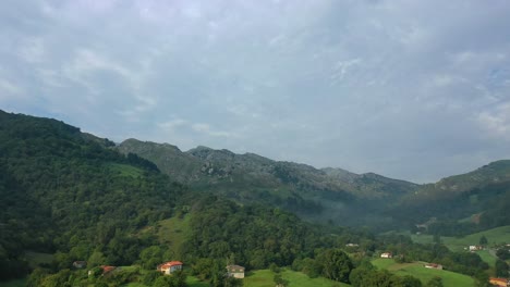 Vuelo-Dando-Un-Giro-En-Un-Valle-En-Verano-Con-Un-Pueblo-Y-Casas-De-Campo-En-Un-Entorno-Verde-Lleno-De-Prados-Y-Bosques-De-Hayas-Y-Robles-Con-Un-Cielo-Nublado-En-Cantabria-España