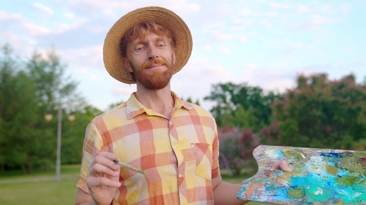 Artista con i capelli rossi con tavolozza e pennello che sorride in un parco