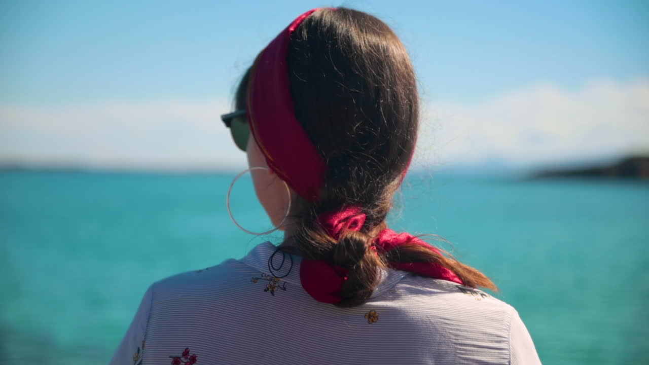 Brunette with fuschia hairband and braid, looking at turquoise ocean, close-up from behind.