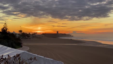 Einsamer-Trator,-Der-Am-Strand-Mit-Eingeschaltetem-Licht-Arbeitet,-Mit-Dramatischem-Sonnenuntergang-Am-Leeren-Strand-Im-Hintergrund,-Randstraße,-Carcavelos