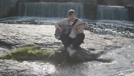 young man playing guitar sitting on the bank of a mountain river on a background of rocks. concept of freedom relaxation. place