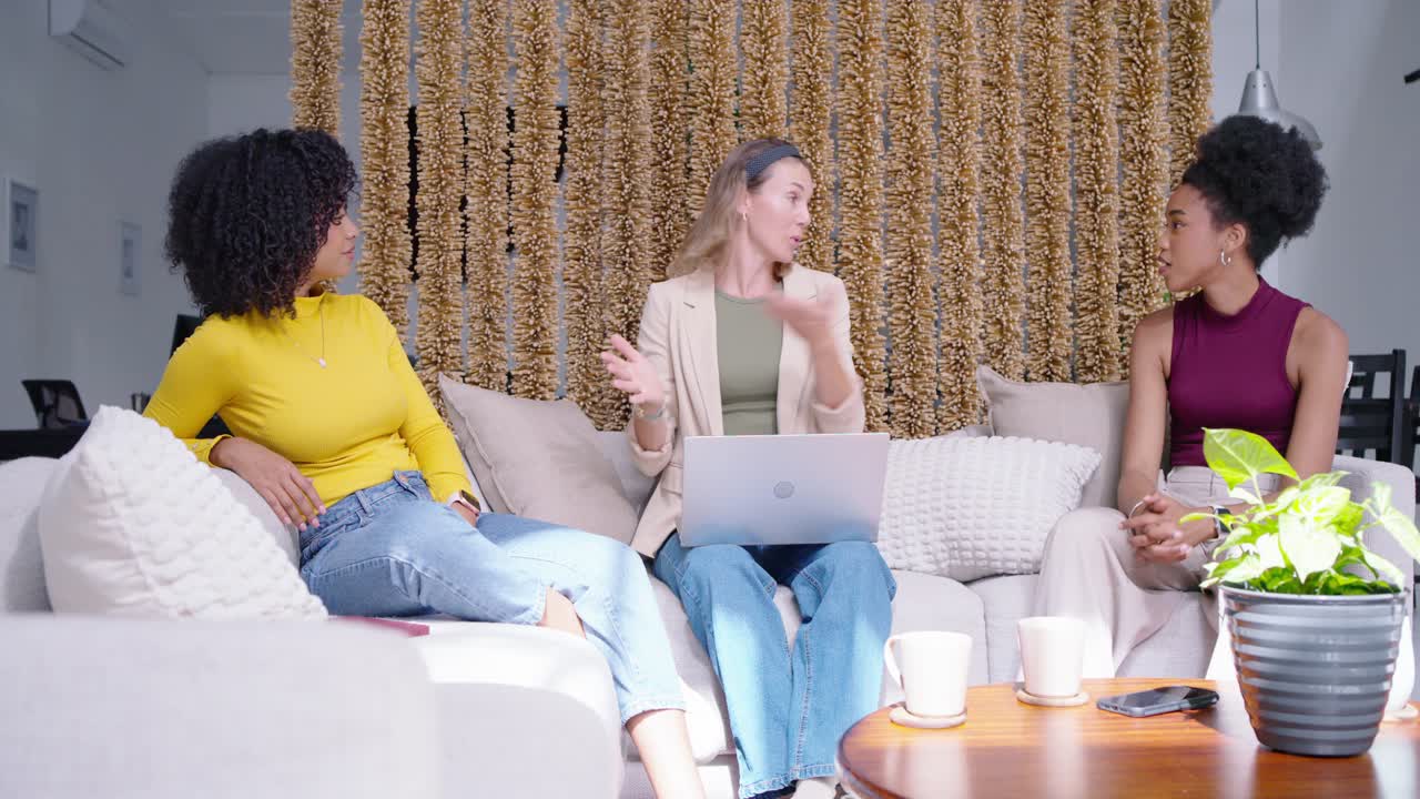 Diverse female coworkers sitting on couch center woman pointing at laptop and mugs explaining plan