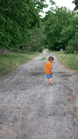 little barefoot kid in orange t-shirt and blue shorts runs by the ground road. toddler spends time outdoors in summer. vertical video.
