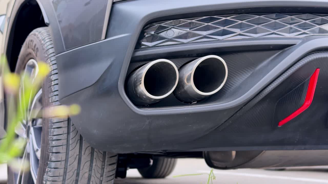Primo piano da angolo basso di doppi terminali di scarico cromati su un diffusore posteriore in fibra di carbonio di un SUV di lusso nero, con cerchio in lega e piante verdi sfocate in primo piano