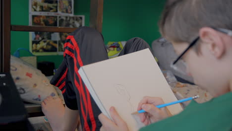 over the shoulder view of a young boy at his desk in his bedroom drawing with a pencil in his sketchbook