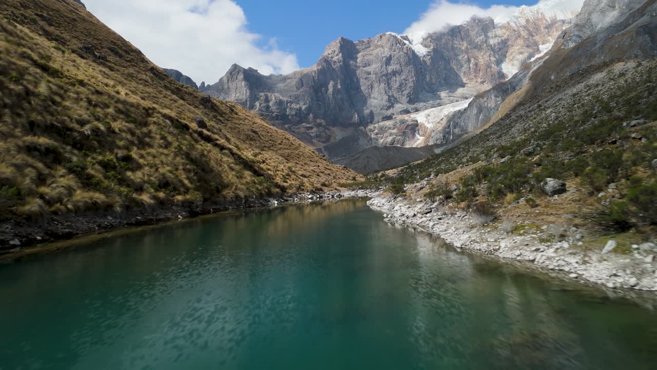 Aerial shot flies forward closely above a stunning turquoise glacial lake, flanked by rugged cliffs and snowy Cordillera Huayhuash peaks in Peru