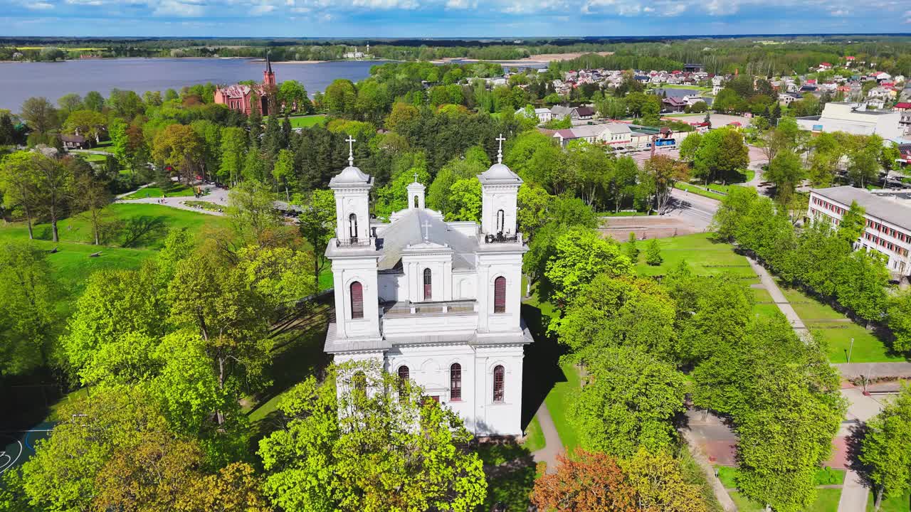 A white church with twin towers stands in the center of a lush park. The aerial view reveals vibrant greenery, footpaths, nearby buildings, and a scenic river horizon.