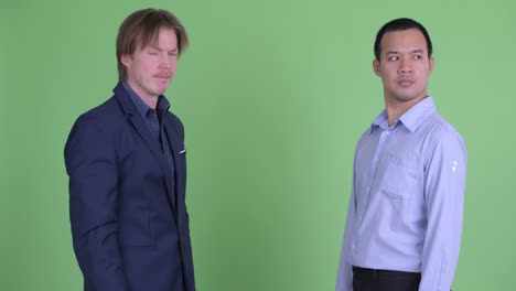 two businessman looking angry and disappointed while having handshake against green studio background