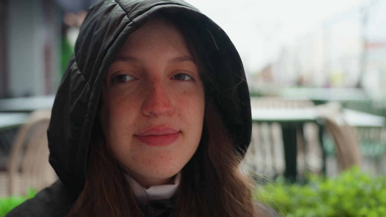 Giovane donna caucasica che sorseggia un caffè all'aperto, cappotto con cappuccio, occhi chiusi, inspira il vapore, terrazza cittadina sullo sfondo, pausa durante la passeggiata, espressione riflessiva, fredda mattina nuvolosa, ritratto spontaneo