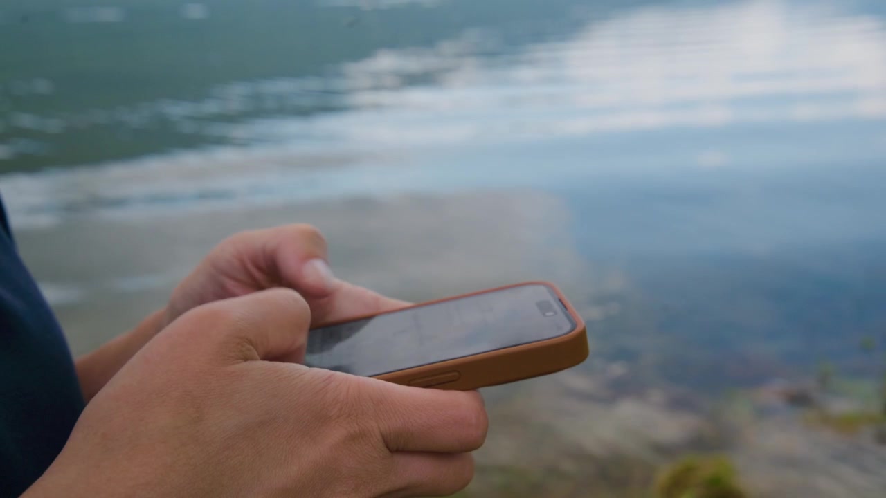 Mano tiene il telefono vicino all'acqua. Persona controlla messaggi o scatta foto. Momento calmo e concentrato in riva al lago. Ottimo per contenuti di viaggio, tecnologia o natura. Adatto per annunci, blog o social media