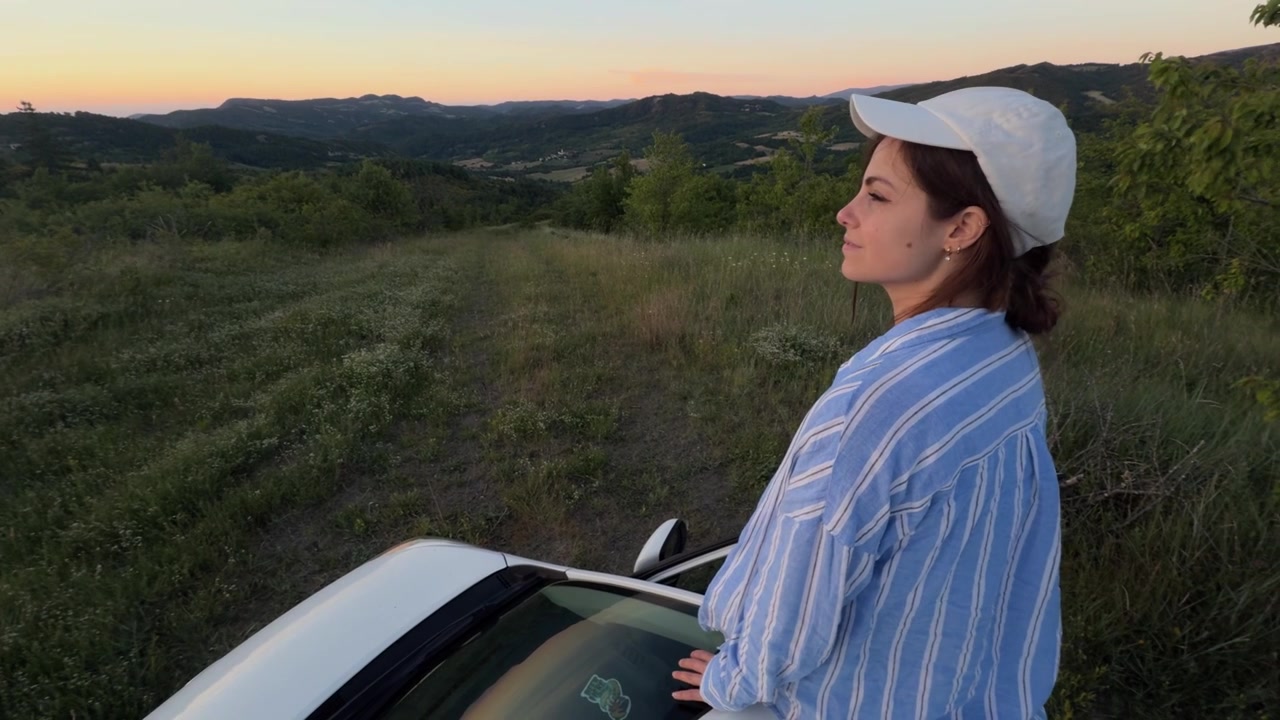 Young thoughtful woman standing near her car while on a road trip. The beautiful female traveler enjoys the beautiful scenic sunset view