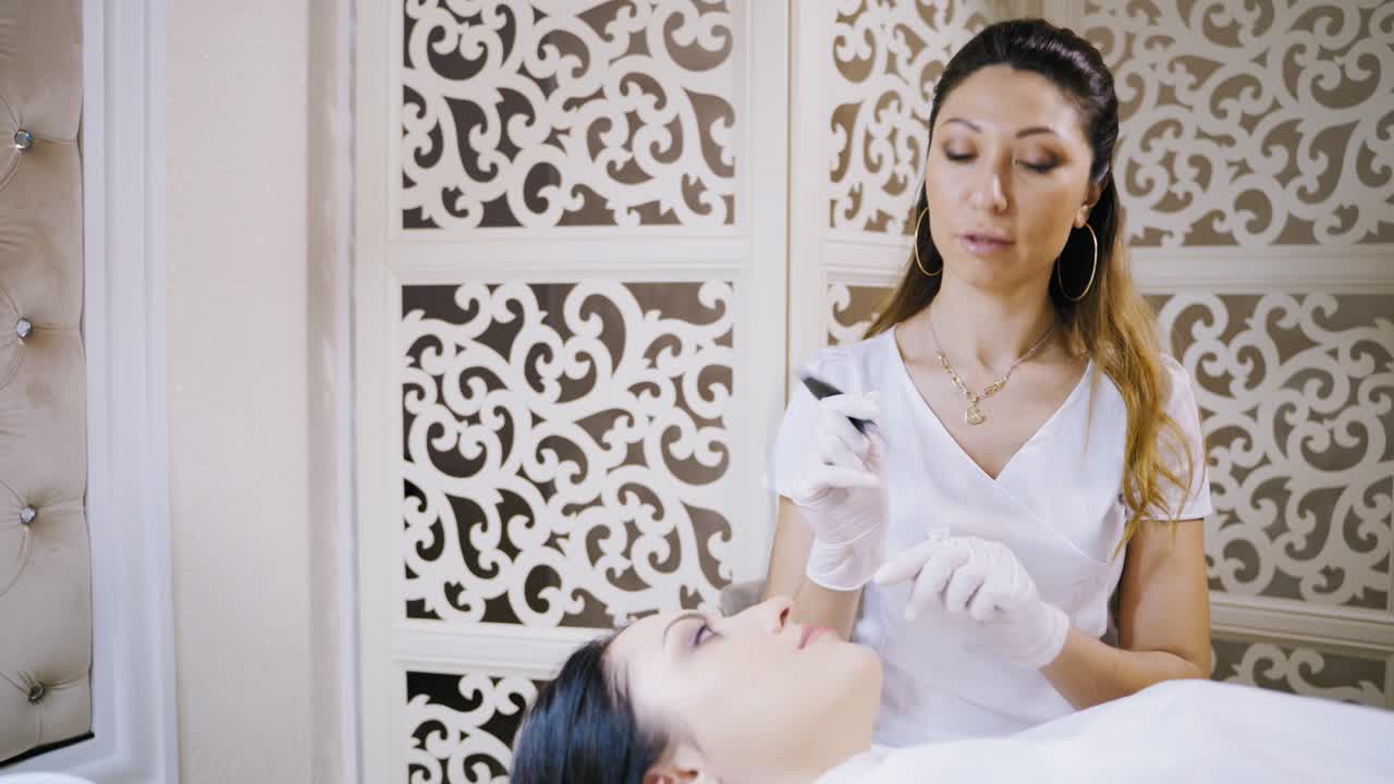 Procedure of permanent make-up for lips of attractive woman in a beauty salon. Pretty young cosmetologist is doing pink tattoo on lips by special machine.