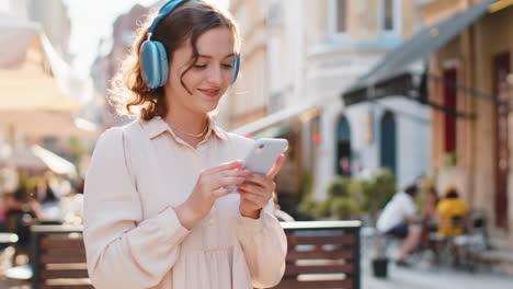 Happy-young-woman-in-wireless-headphones-choosing,-listening-music-dancing-outdoors-city-street