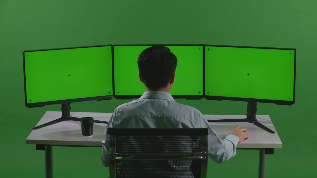 녹색 스크린 스튜디오에서 모의 여러 컴퓨터 모니터와 함께 작업하는 동안 스트레칭하는 피곤한 아시아 남자의 뒷면