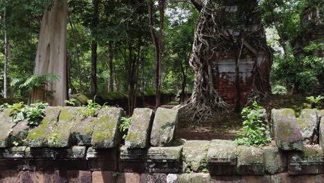 Flight-over-stone-wall-to-overgrown-temple-tower-at-Koh-Ker,-Cambodia