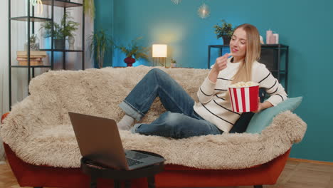 mujer caucásica disfrutando de una película en el sofá comiendo palomitas de maíz riendo viendo la computadora portátil en el interior sonriendo feliz