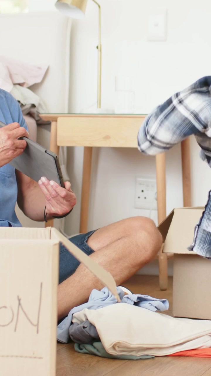 Vertical video: Checking tablet, man in blue shirt folding clothes at home and packing donation box