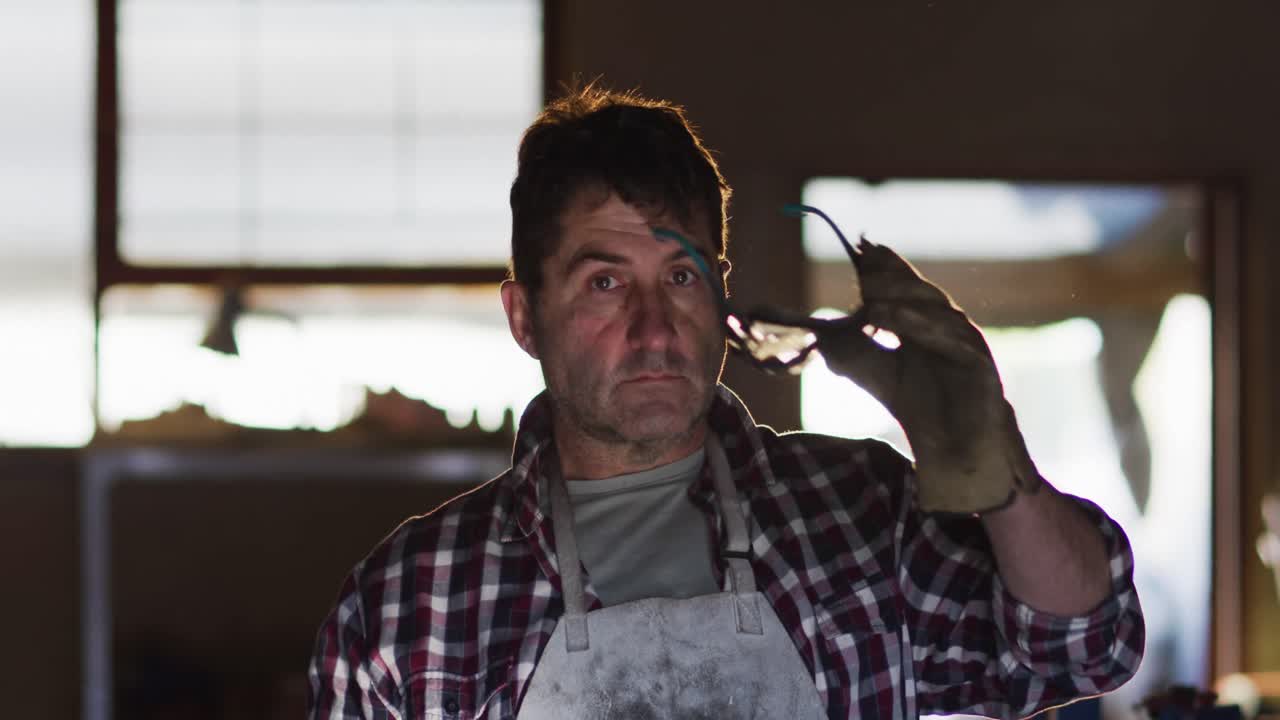 작업실에서 칼을 만드는 백인 남성 안경을 벗고 카메라를 쳐다보고 미소 짓습니다.