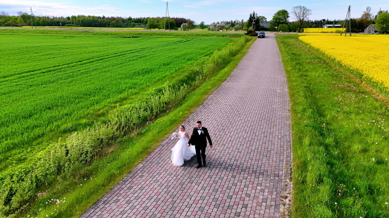 Bride in flowing gown and groom in dark suit walk hand in hand along a cobblestone country road, framed by vast golden fields and lush spring greenery