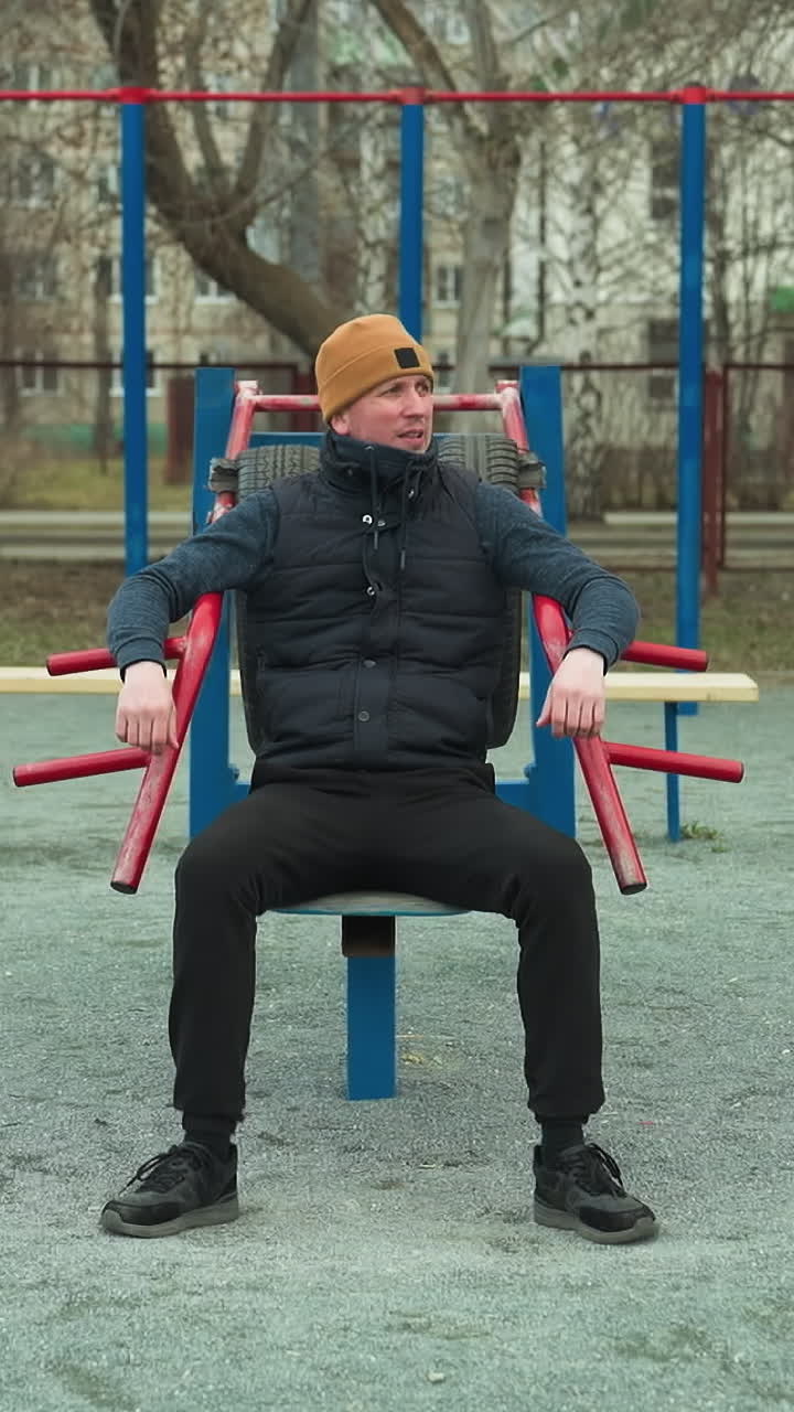 A coach adjusts himself on outdoor workout equipment, while two boys play football in the background, one boy in a gray sweater kicks the ball away from the boy in black