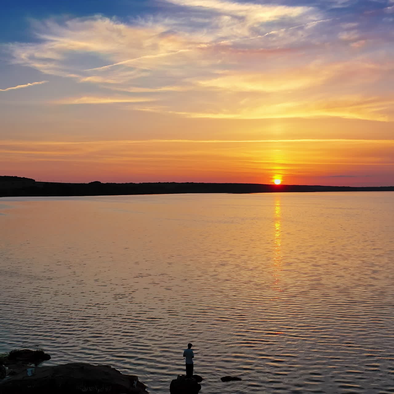 Flying over the evening river and some people standing on a stone in water. Golden sun setting on the natural water background. Silhouette of a fisherman at sunset.
