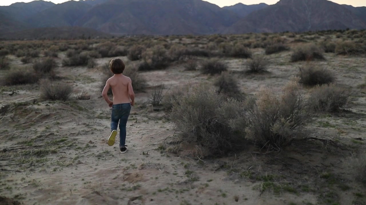 Giovane ragazzo in jeans blu a torso nudo che corre attraverso il deserto con montagne sullo sfondo al tramonto.