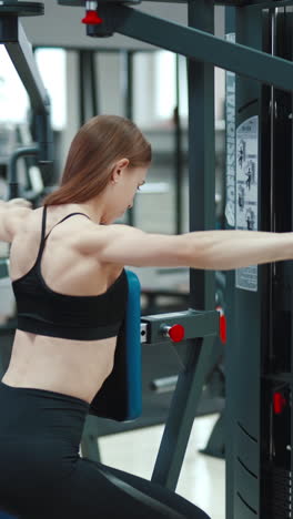 a woman with a sporty figure is sitting and doing an exercise to strengthen the back muscles in the gym. blurred background vertical video