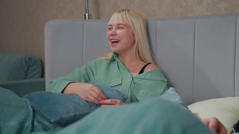 blonde girl in green shirt smiling warmly while lying on bed, enjoying lighthearted conversation with friend partially visible in cozy bedroom