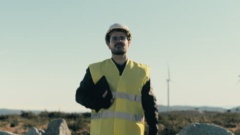 a skilled caucasian male engineer in safety gear inspects wind turbines while walking in a field of renewable energy generators on a sunny day, emphasizing the significance of renewable energy