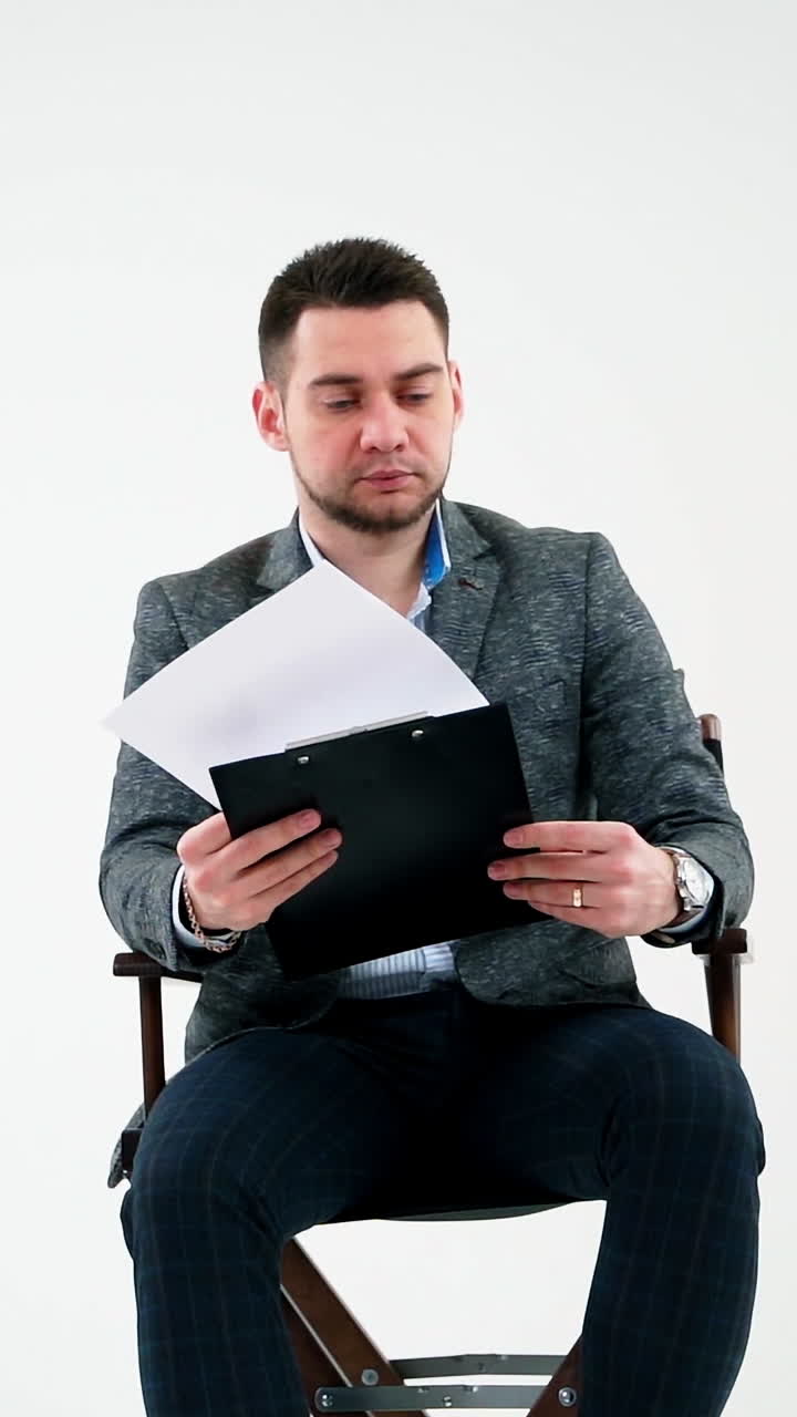Serious businessman holding a folder and reading documents. Paper works flying over the handsome man in grey suit isolated on white studio background. Vertical video